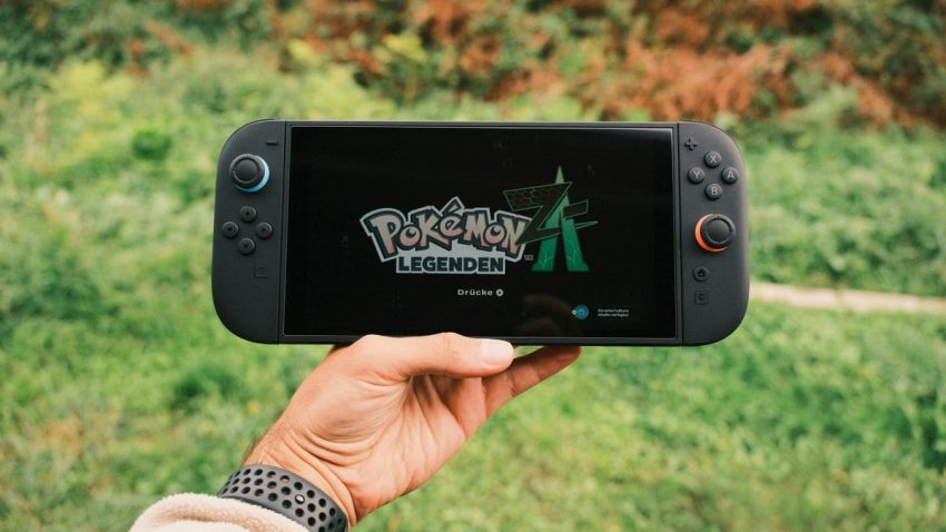A hand holding a handheld gaming console displaying the Pokémon Legends game screen outdoors.
